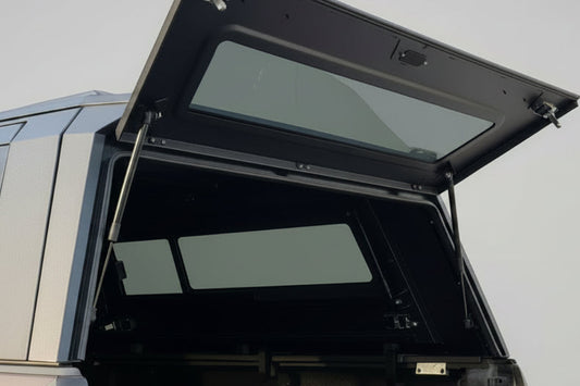 A black hardtop canopy tent mounted on the rear of a vehicle, shown with its upper door panel open in an outdoor residential setting.