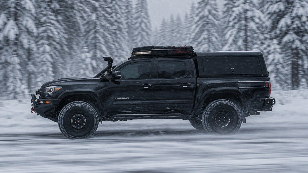 A black Toyota Tacoma pickup truck with a hardtop canopy and roof rack drives through deep snow in a forested winter setting.