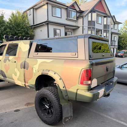 A lifted Ram pickup truck with a camouflage wrap in olive green, tan, and brown, parked on a street in front of multi-story residential buildings.