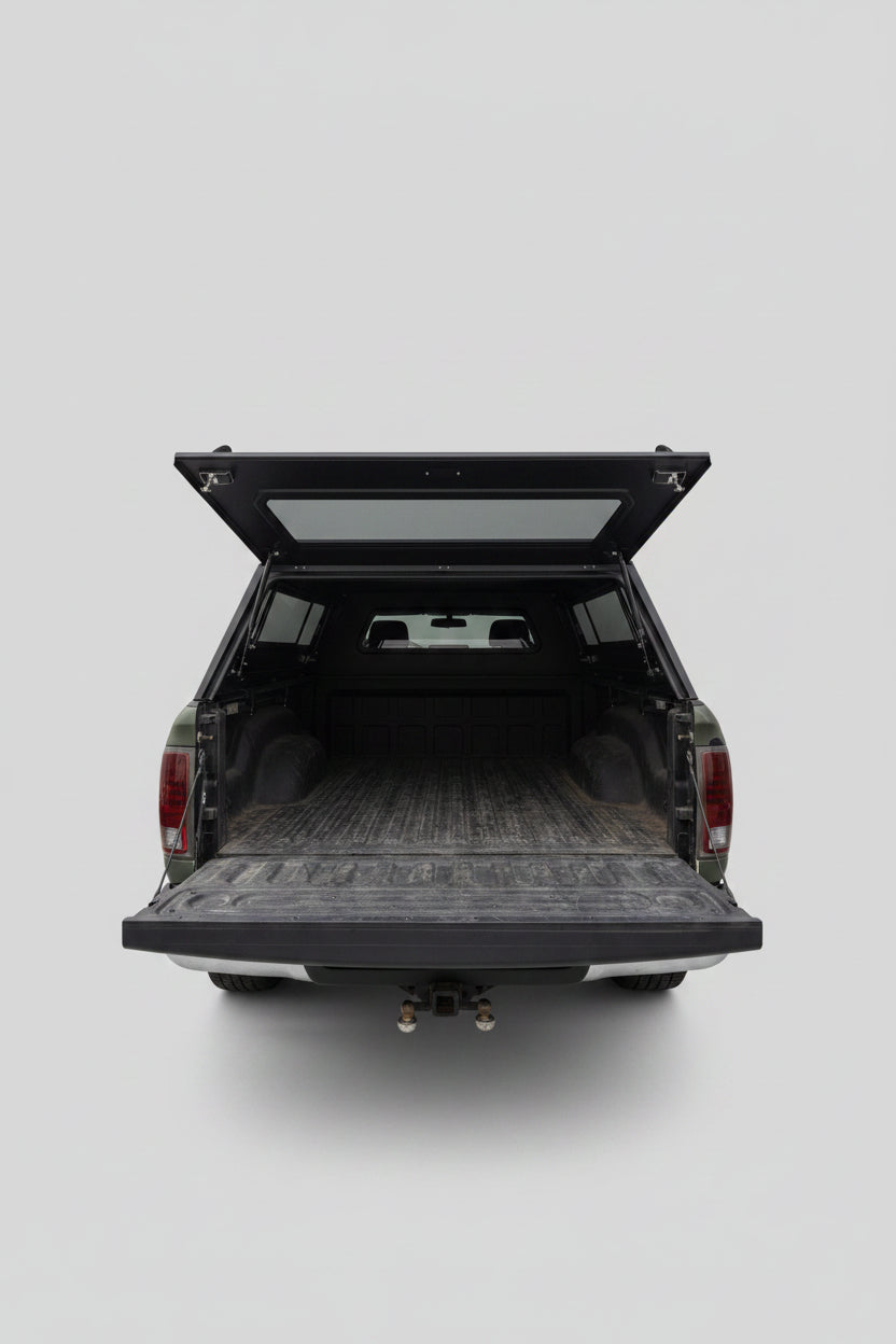 The back of a dark pickup truck with the tailgate and hatch open, showing a cluttered cargo bed with various items inside, parked in a residential driveway with houses and a blue sky in the background.