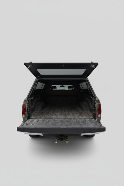 The back of a dark pickup truck with the tailgate and hatch open, showing a cluttered cargo bed with various items inside, parked in a residential driveway with houses and a blue sky in the background.
