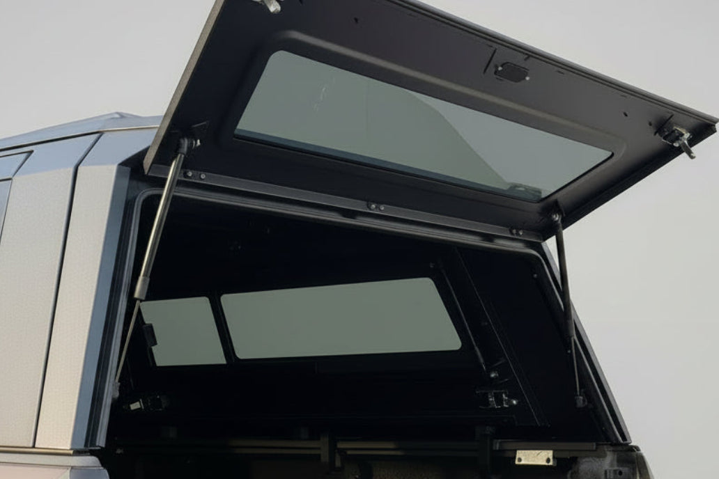 A black hardtop canopy tent mounted on the rear of a vehicle, shown with its upper door panel open in an outdoor residential setting.