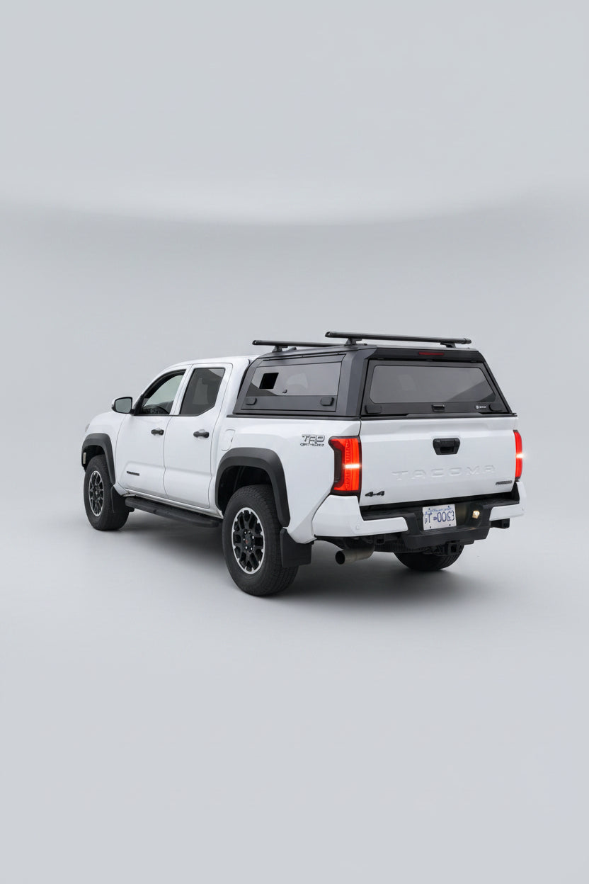 A white Toyota Tacoma pickup truck parked outdoors in a residential driveway, viewed from the rear three-quarter angle. The truck has a black hardtop canopy over the bed, black wheels, and red taillights illuminated.