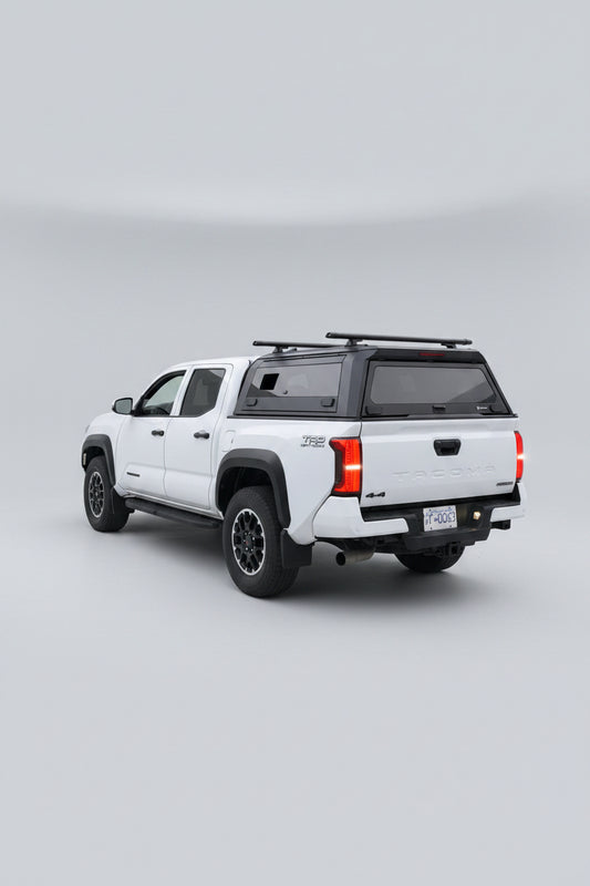 A white Toyota Tacoma pickup truck parked outdoors in a residential driveway, viewed from the rear three-quarter angle. The truck has a black hardtop canopy over the bed, black wheels, and red taillights illuminated.